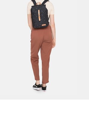 Personne portant un pantalon couleur rouille, des baskets noir et blanc, et un sac à dos noir avec des détails de sangle marron, se tenant de dos.