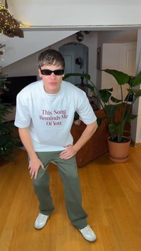 Λευκό T-shirt με κόκκινο κείμενο που γράφει «This Song Reminds Me Of You», παντελόνι χακί, μαύρα γυαλιά ηλίου και λευκά αθλητικά παπούτσια σε ξύλινο πάτωμα.