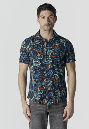 Hombre con camisa de manga corta abotonada con patrones tropicales de hojas y pájaros en azul, verde y naranja, combinada con jeans grises.