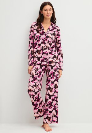 Vrouw staat blootsvoets en draagt een bijpassend shirt met lange mouwen en wijde broek met roze en paarse bloemenprint op zwarte achtergrond.