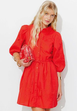 Femme blonde portant une robe rouge boutonnée avec des manches bouffantes, tenant une pochette rouge à motif, accessoirisée avec des bracelets dorés et des boucles d’oreilles créoles.