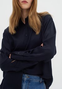 Hajózási kék, hosszú ujjú, gombos ing sima anyagból, gallérral, elülső gombokkal és gombos részletekkel ellátott mandzsettával, farmernadrággal viselve.