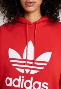 Kvinna som bär en röd Adidas-hoodie med vitt trefoil-logotyp och dragsnören, som visar hennes läppar, hakan och lockigt hår.