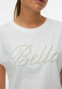 Sieviete, kas valkā baltu T-kreklu, uz kura krūtīm ir rakstīts "Bella" dekoratīvu pērļu pērlītēs.