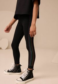 Schwarze Leggings mit einem bunten Strassstreifen an der Seite, kombiniert mit schwarzen High-Top-Sneakern mit weißen Sohlen und Schnürsenkeln.