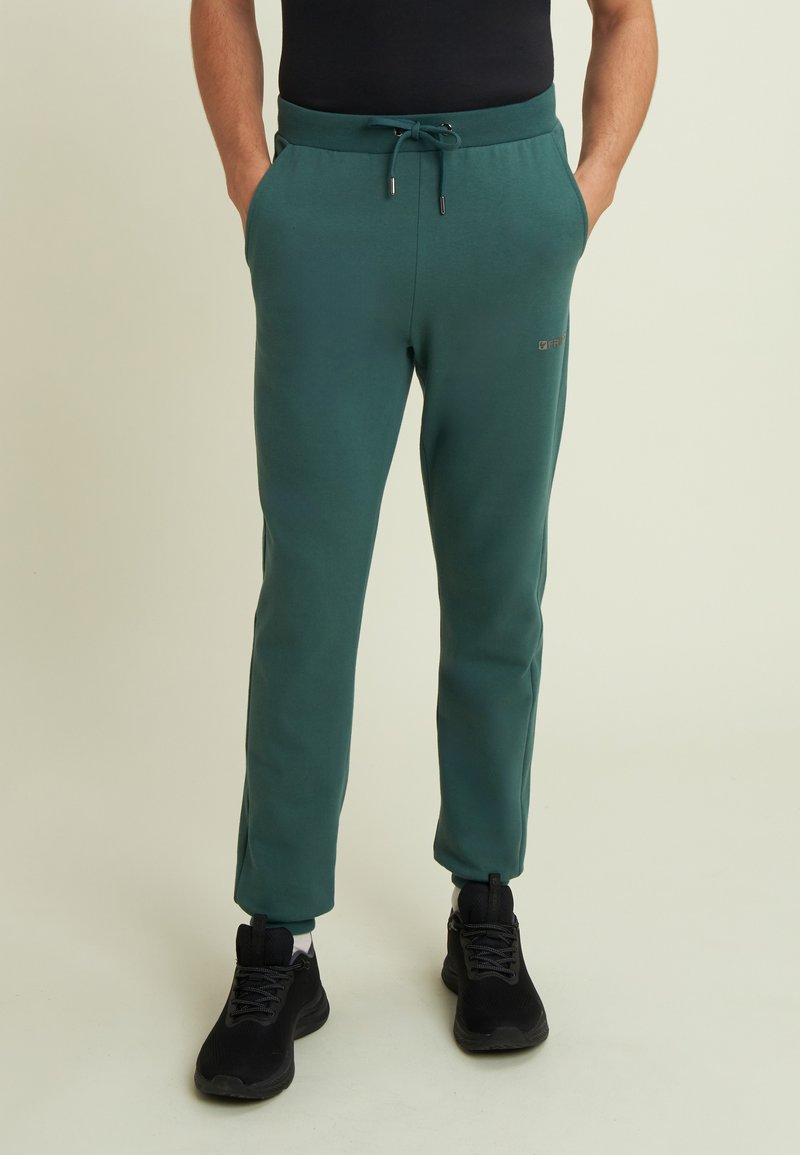 Hombre vistiendo pantalones jogger teal con cordón y zapatillas negras, de pie con las manos en los bolsillos contra un fondo sencillo.