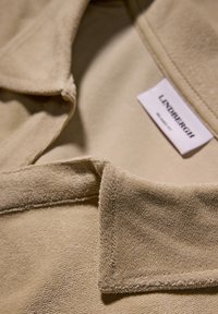 Beige Stoff mit Kragen, weiche Textur mit einem plüschigen finish, versehen mit einem Label, auf dem "LINDREGH" in schwarzer Schrift steht. Die Kanten sind sauber genäht.