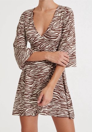 Femme portant une robe portefeuille à motif zèbre marron et crème avec des manches trois-quarts, mains croisées à la taille, sur un fond clair uni.