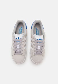 Grå sneakers i mocka med vita detaljer, tre vita ränder och blå logotyp på tunga och häl. Rundad tå och snörning.