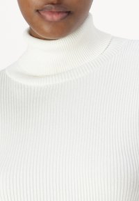 Pull à col roulé côtelé blanc avec un design près du corps, présentant un col haut et une texture douce, mettant en valeur les lignes horizontales et les détails.