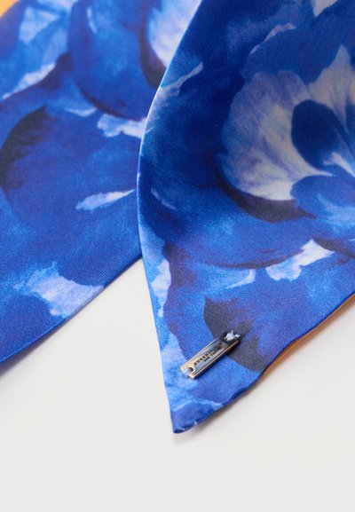 Μπλε ύφασμα με λουλουδάτο μοτίβο και μεταλλική ετικέτα με λογότυπο Tag Heuer προσαρτημένη κοντά στην αιχμηρή άκρη σε λευκό φόντο.