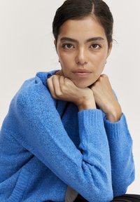 Blå texturerad tröja med ribbade mudd. Tyget ser mjukt ut och har en löst stickad design. Modellen har händerna vilsamt under hakan.