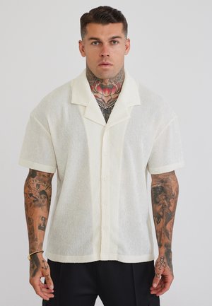 Mann mit Hals- und Arm-Tattoos, trägt ein weißes, kurzärmeliges, strukturiertes Hemd mit Knöpfen und schwarze Hose vor schlichtem Hintergrund.