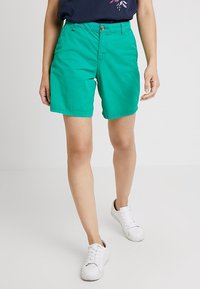 Shorts de algodón verdes con un corte regular, cremallera y un solo botón en la parte frontal. Combinados con zapatillas blancas sobre un fondo liso.