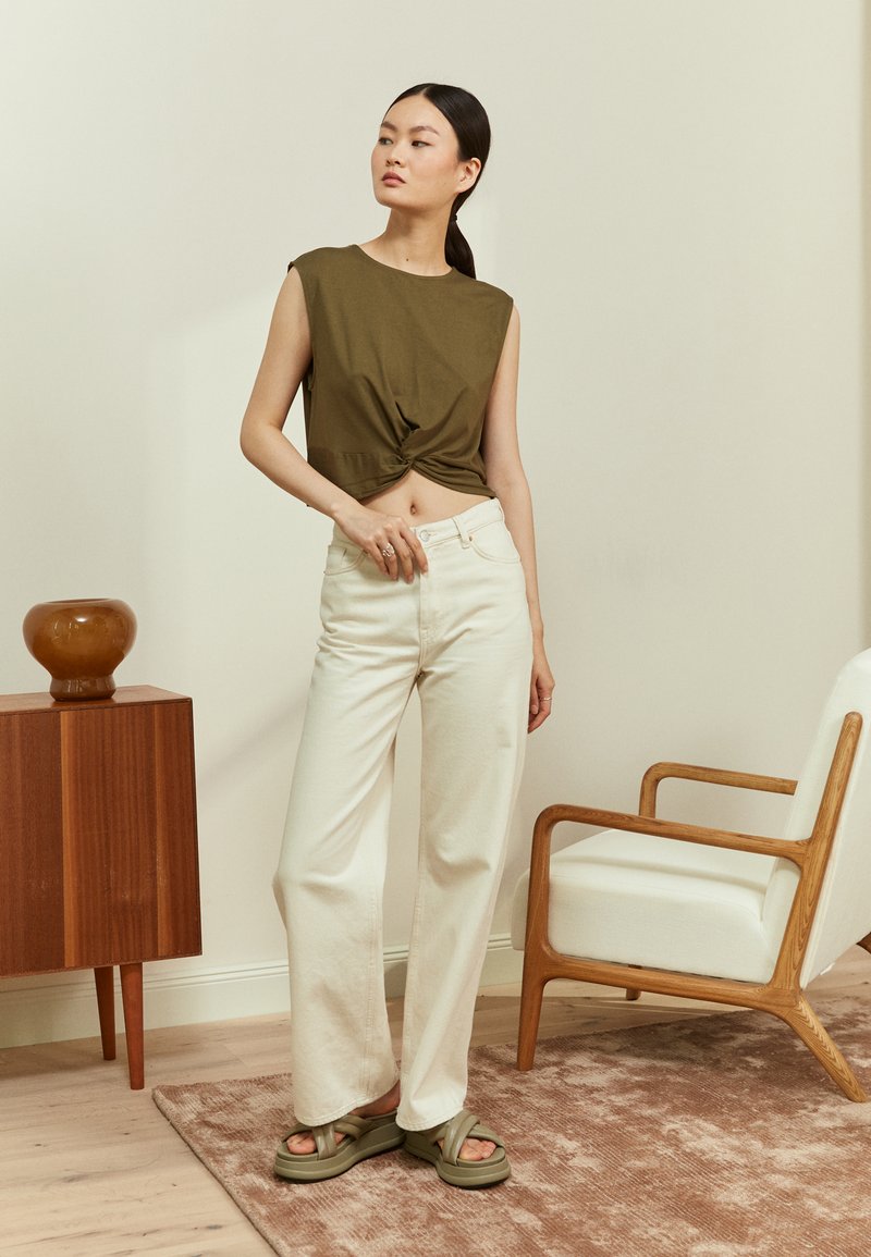 Top cropped verde oliva com detalhe de nó, combinado com calças largas creme e sandálias de plataforma verdes, situado num espaço interior moderno.