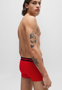 Boxer-briefs rouges en coton avec un élastique noir et rouge portant le logo "HUGO". Le modèle affiche des tatouages sur le bras.