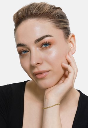Gros plan sur une personne à la peau claire et aux yeux bleus, portant un haut noir. Elle a un délicat bracelet en or à son poignet.