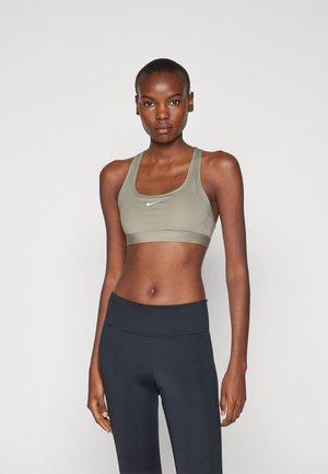 Sujetador deportivo verde oliva con diseño de espalda al descubierto y logo de Nike en blanco, combinado con leggings negros de cintura alta. Textura suave y ajustada.