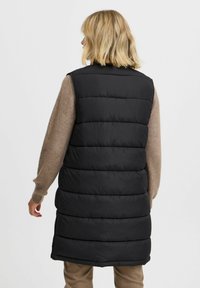 Doudoune noire à conception longue, dotée de sections quiltées horizontales et d'un col montant, portée sur un pull beige clair.