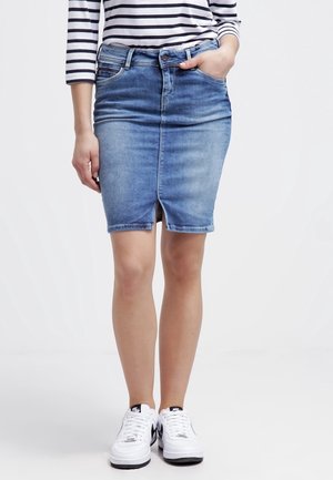 Frau trägt einen blauen Jeansrock mit einem vorderen Schlitz, ein schwarz-weiß gestreiftes Oberteil und weiße Nike-Sneaker, steht vor einem weißen Hintergrund.