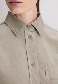 Light beige collared shirt made of soft fabric featuring a button-up front with two buttons visible and a chest pocket.
