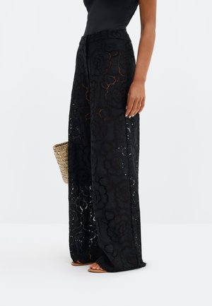 Sieviete, valkājot melnas plašas mežģīņu bikses ar ziedu rakstu, turot austu salmu somu un stāvot basām kājām uz baltas grīdas.