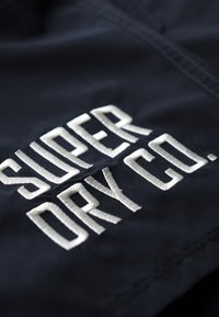 Tejido marino con prominente texto bordado en blanco "SUPER DRY CO." Presenta una textura suave y una costura ordenada a lo largo de los bordes.
