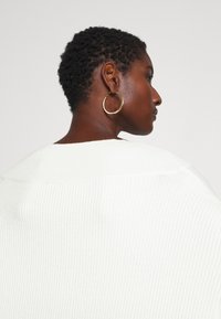 Ribbad vit tröja som hänger över axlarna; modellen har kort hår och bär en stor guldring i örat. Neutral bakgrund.