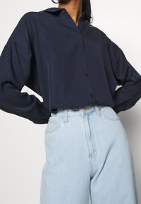 Námořnická modrá košile na knoflíky s uvolněným střihem, dlouhými rukávy a límečkem, spárovaná se světle modrými džíny s vysokým pasem.