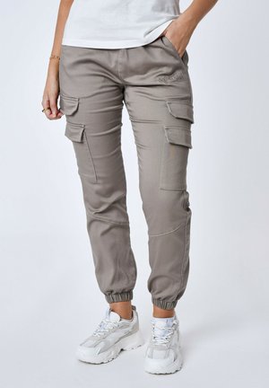 Pantalons cargo en tissu beige clair avec des poignets élastiques, plusieurs poches latérales et des détails de couture ton sur ton. Associés à des baskets de sport blanches.
