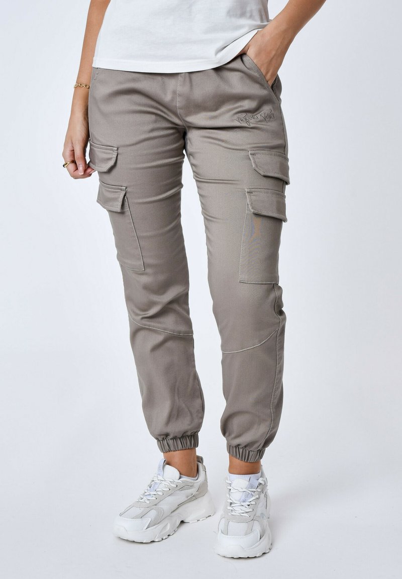 Pantaloni cargo in tessuto beige chiaro con polsini elastici, più tasche laterali e dettagli di cucitura tono su tono. Abbinati a scarpe da ginnastica bianche.