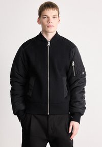 Casaco bomber preto com textura canelada no corpo, mangas acolchoadas, fecho de correr e bolsos laterais. Apresenta um pequeno logótipo na manga.