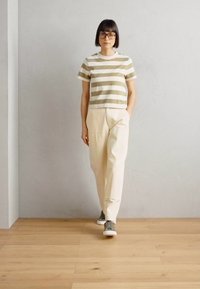 Modelo veste uma t-shirt de manga curta às riscas em oliva e creme, combinada com calças creme e ténis cinzentos, de pé contra um fundo neutro.