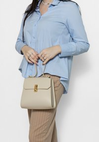 Borsa strutturata beige con una chiusura dorata, texture liscia e forma rettangolare, tenuta da una persona in camicia azzurro chiaro e pantaloni a fantasia.