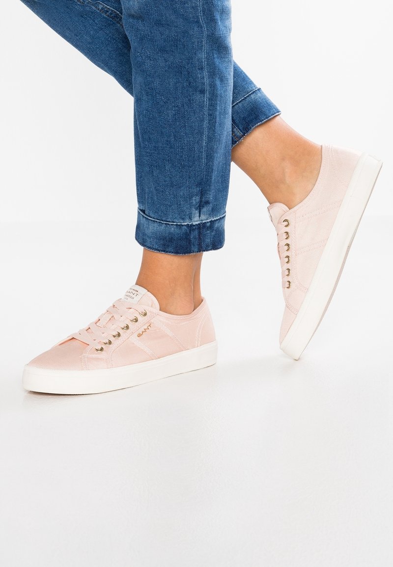 Une personne portant des baskets en toile roses claires et un jean bleu retroussé devant un fond blanc uni.