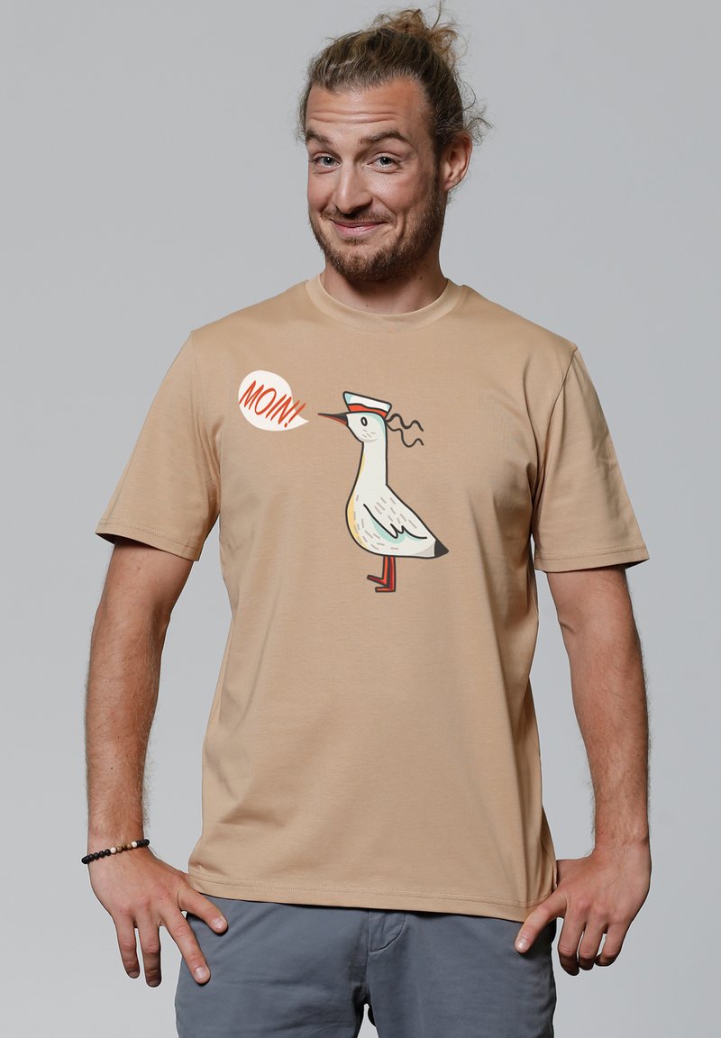 Beige t-shirt met korte mouwen, met daarop een cartoonvogel met een hoed en het woord "MOIN!" in een spraakballon. Zacht materiaal en een relaxte pasvorm.