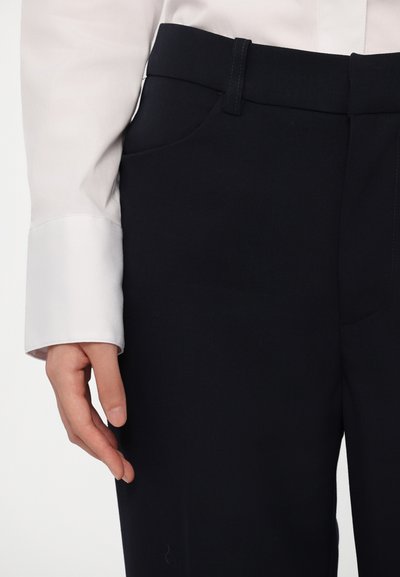 Zwarte op maat gemaakte broek met een vlakke tailleband, zijzakken en een gladde textuur; getoond met een witte shirtmouw en een hand die ernaast rust.