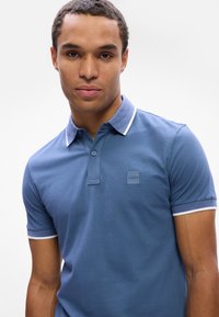 Polo bleu avec un col et des bords de manches blancs contrastants, arborant un discret logo brodé sur la poitrine. Texture en coton lisse.