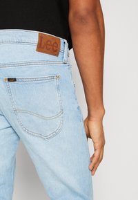 Höger arm och hand bredvid ljusblå Lee jeans med bakfickan och brunt Lee-logotypmärke, på en person klädd i svart topp mot en enkel bakgrund.