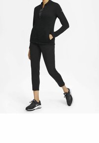 Person som bär en svart zip-up jacka, svarta avkortade byxor och svarta Nike-sneakers, står med en hand i fickan och benet sträckt framåt.