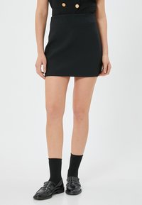 Zwarte mini-jurk met een aansluitende silhouet, gemaakt van gladde stof, gecombineerd met zwarte loafers en sokken, staand tegen een effen witte achtergrond.