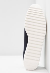 Zool van een zwart-witte sneaker met diepe horizontale groeven, leunend tegen een witte hoekmuur op een witte ondergrond.
