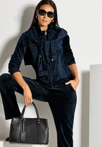 Marineblauer Velours-Trainingsanzug mit strukturierten Details, Reißverschluss und Kapuze; kombiniert mit einer schwarzen Ledertasche mit polierten Beschlägen und Griffen.