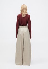 Femme blonde de dos portant un haut rouge foncé à manches longues et un pantalon beige taille haute à jambes larges sur un fond clair uni.