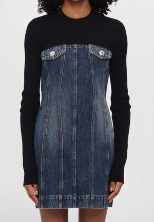 Kobieta w ciemnoniebieskiej, bezramiennej sukience jeansowej na czarnym, dopasowanym, długim rękawie, stoi na tle jednolitego jasnego tła.