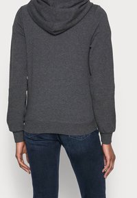 Grijze hoodie gemaakt van zacht materiaal, met een kangoeroezak, ribgebreide manchetten en zoom. Achteraan is een ontspannen pasvorm zichtbaar.