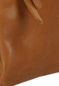 Borsa in pelle di colore marrone chiaro con una texture liscia. Presenta un design semplice e un logo goffrato discreto sul lato.