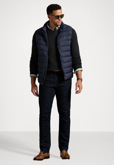 Marineblauwe puffer vest over een zwart kabelgebreid sweater, gecombineerd met donkere jeans en bruine leren schoenen; accesoriseert met een zonnebril.