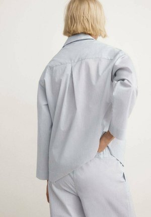 Persona con cabello corto y rubio, vestida con una camisa holgada a rayas finas azul claro y blanca, y pantalones a juego, de pie con la mano en el bolsillo trasero.