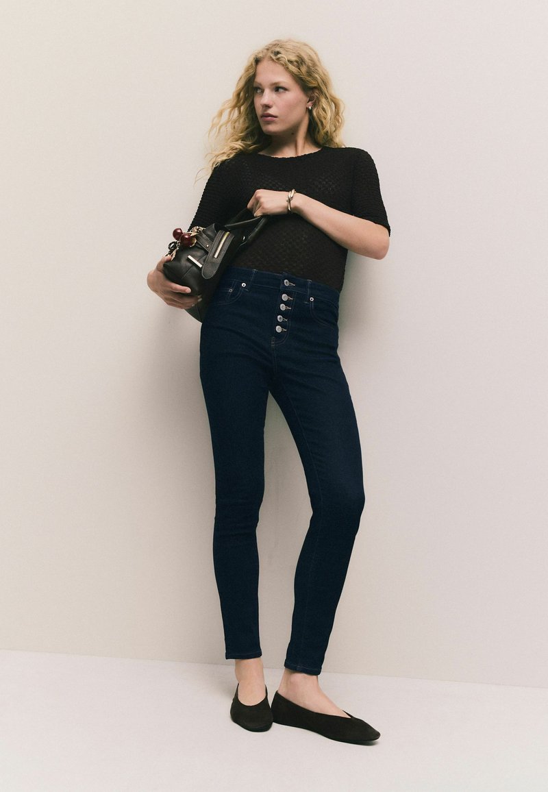 Augsti vidukļa tumši zilie džinsi ar piecām pogām, apvienoti ar teksturētu melnu topu un melniem apaviem, uz neitrāla fona.